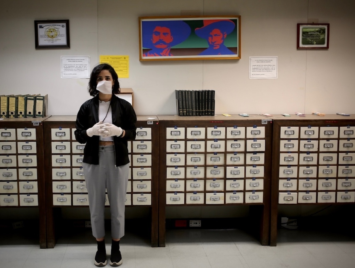 A woman wearing a face mask and gloves stands in front of a wall of library card catalogue drawers.