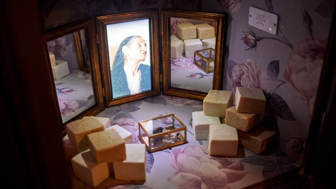 Locker interior housing a triptych mirror with a photo of an elderly woman, piles of soap, and a glass display box.