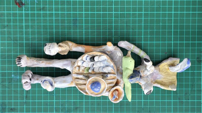 A small clay figurine, part human, part animal, with a green carrot in its beak, lying on a green gridded cutting map.