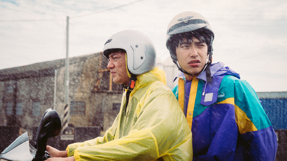 An image of two men riding a moped through an industrial area. It is raining heavily and both wear colourful raincoats.