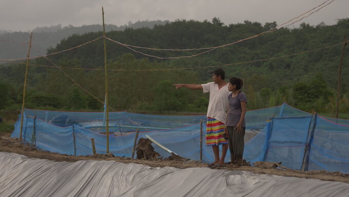 An overcast, wide shot shows a Taiwanese businessman and Burmese worker stood by a newly constructed aquaculture pond.