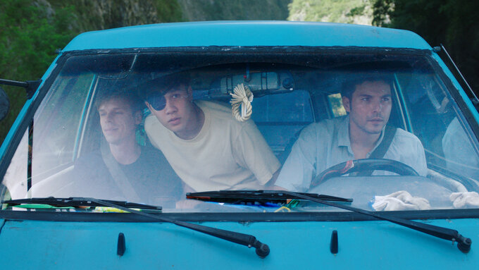 A close-up color shot taken through the front windshield of a blue van, showing 3 young men sat in the front cab.