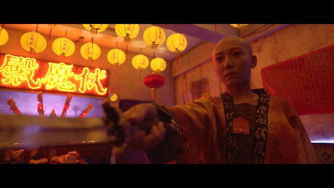 A highly stylised, darkly lit shot in a temple. A bald woman in a robe holds a sword. Lit by neon signs & lanterns.