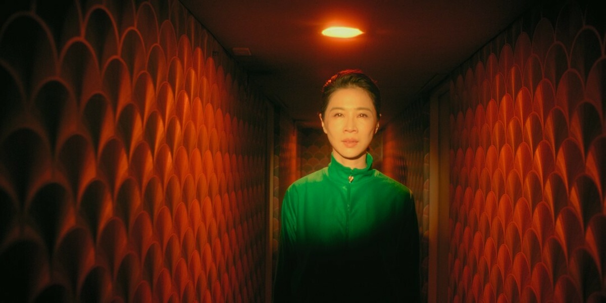 A saturated colour shot of a woman in a bright, high-collared green top stood in the centre of a narrow corridor.