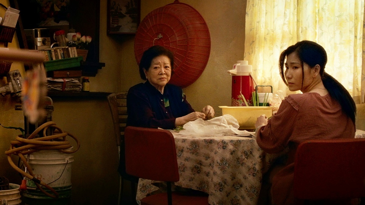 A medium close-up shot in a warm, cluttered home interior. Two women sit at a table covered with a floral cloth.