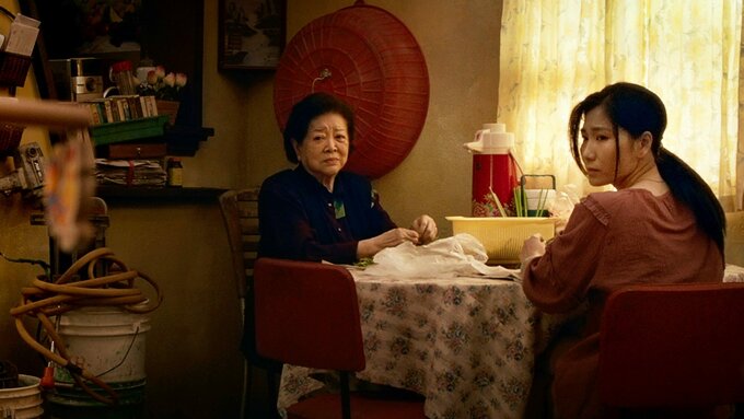 A medium close-up shot in a warm, cluttered home interior. Two women sit at a table covered with a floral cloth.