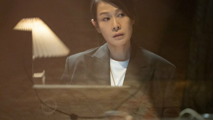 A warm, softly lit, close-up shot of a woman in a grey blazer. She is sat at a desk, with a contemplative expression.