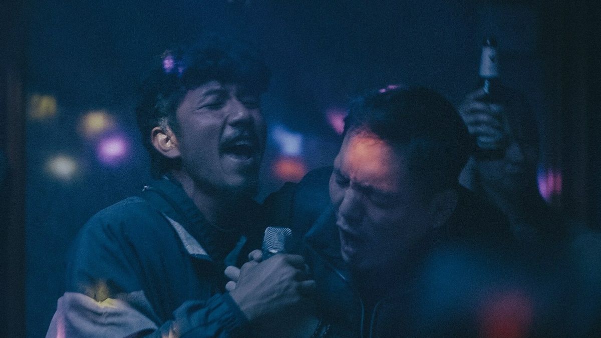 A darkly lit, grainy color shot of two men passionately singing karaoke.