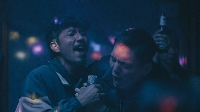 A darkly lit, grainy color shot of two men passionately singing karaoke.