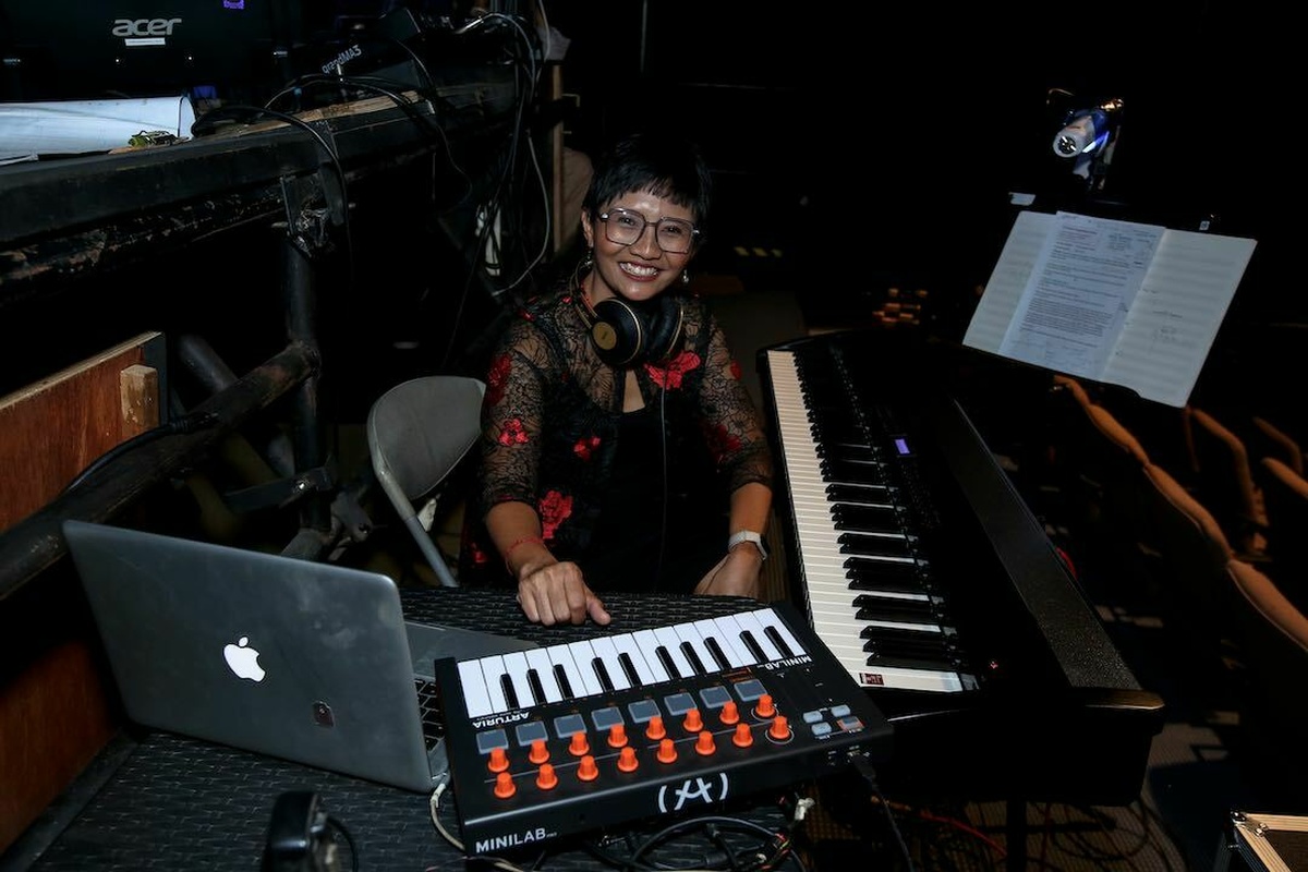 A woman amidst keyboards and music equipment, smiling.