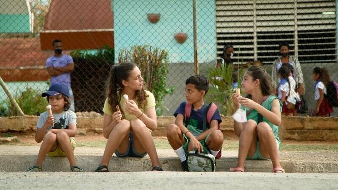4 people sit on a pavement eating icecreams. Three of them are children.
