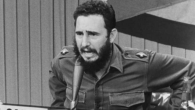 A man with a beard and a Cuban flag in the background speaks into a microphone