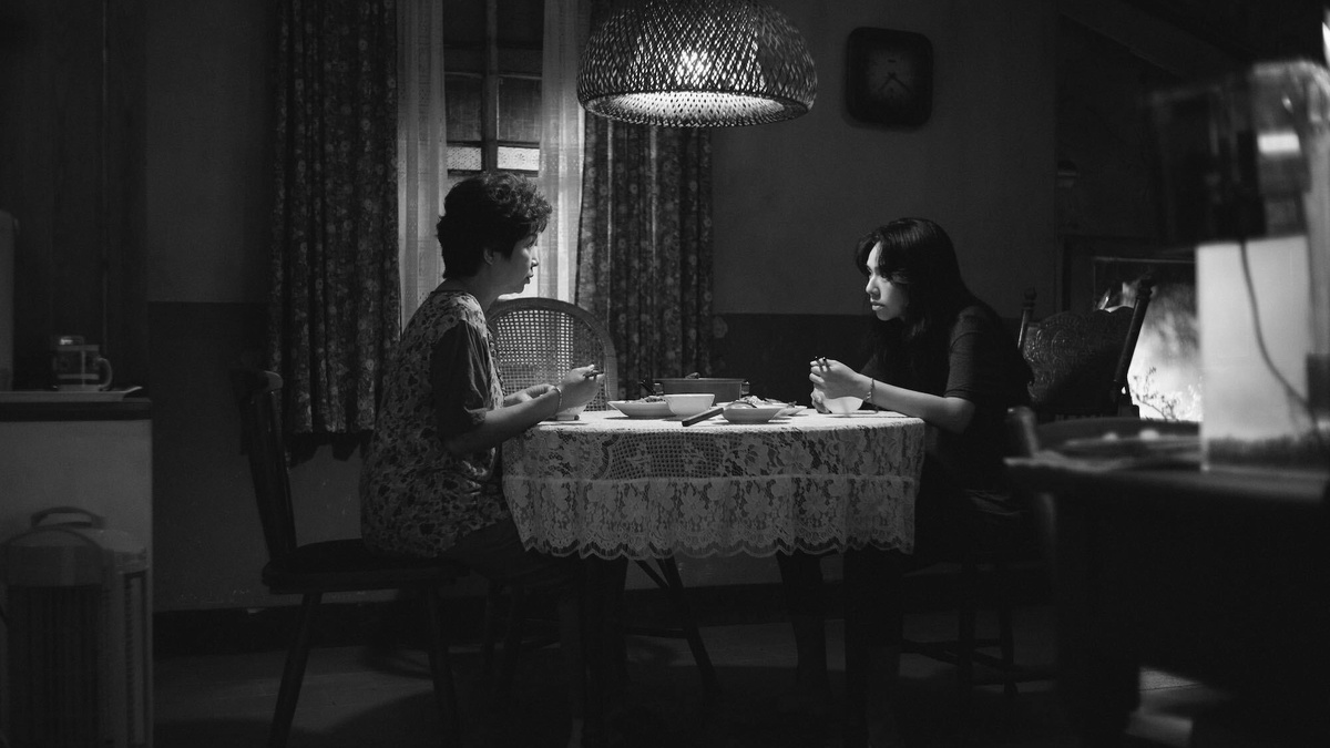 A tense, black and white interior shot. 2 women 1 older and 1 younger sit facing each other at a kitchen table.