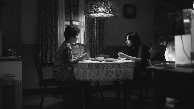 A tense, black and white interior shot. 2 women 1 older and 1 younger sit facing each other at a kitchen table.