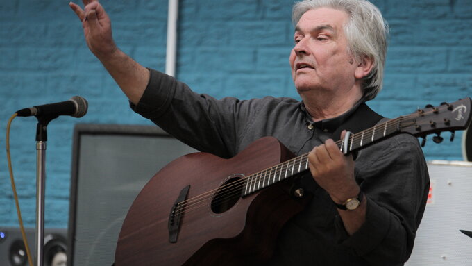 A light skinned man with grey hair holding a guitar. His hand is raised in an 'OK' gesture.