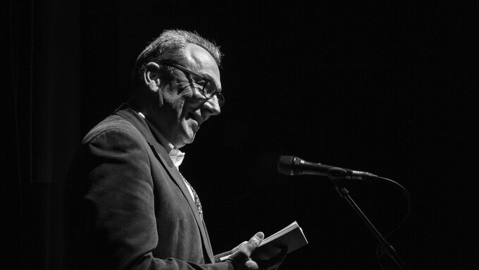 A black and white image of a man reading from a book of poetry.