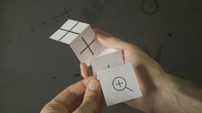 Two hands unfold a piece of paper. On the front is printed the "zoom" or magnifying glass symbol.
