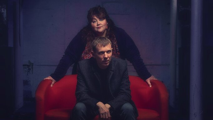 A man sat in an red arm chair with a woman standing over him. Both have pale skin and dark hair and wear dark clothing.