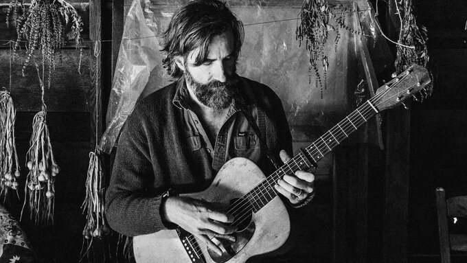 A black and white photo of a light skinned bearded man playing guitar in a log cabin.