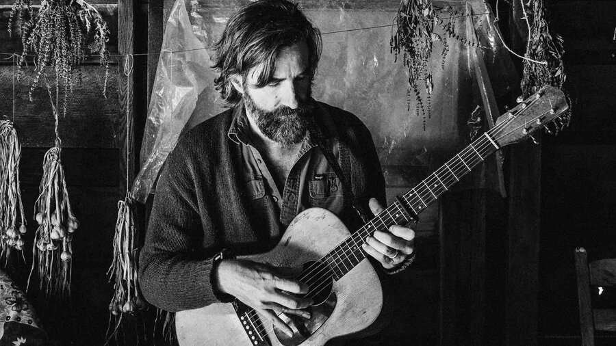 A black and white photo of a light skinned bearded man playing guitar in a log cabin.