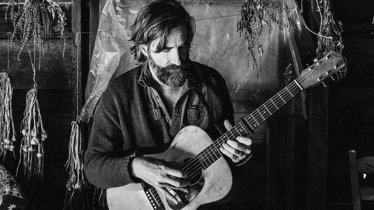 A black and white photo of a light skinned bearded man playing guitar in a log cabin.