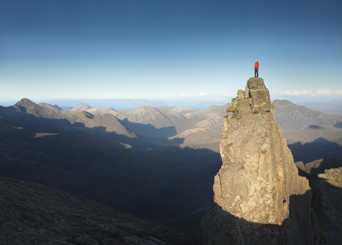 A man in climbing gear stood at the top of a large rocky outcrop, you can see rolling hills and blue sky behind him.