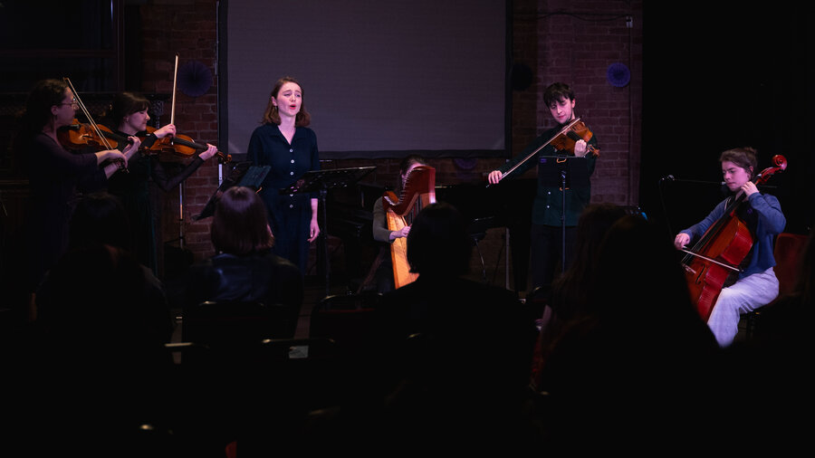 A woman singing alongside a band. Including a violinists, chellists, and a harp player.