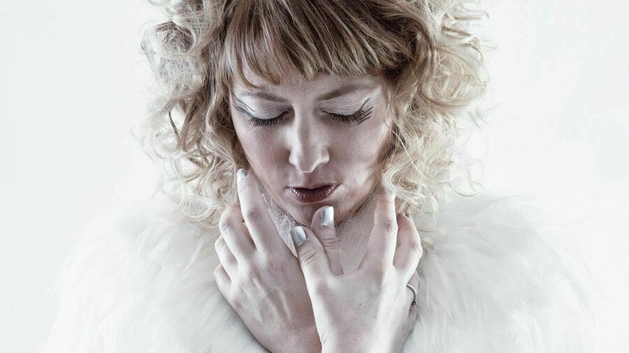 A performer stands against a white background with eyes closed, wearing a blue dress with a white feathered collar.