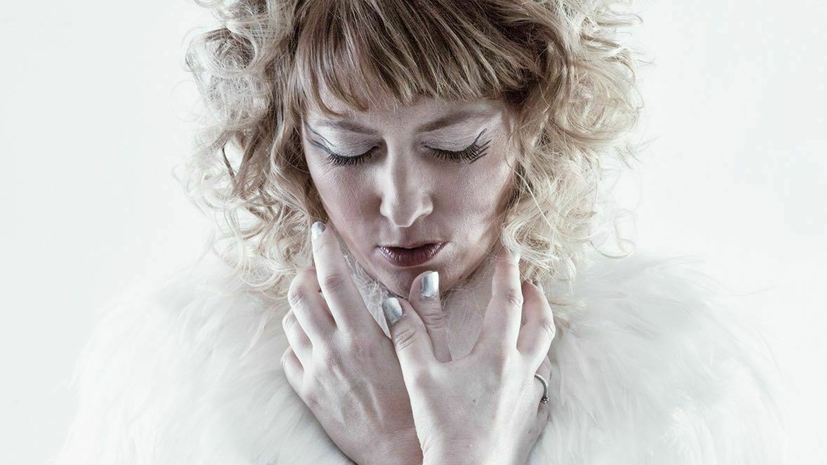 A performer stands against a white background with eyes closed, wearing a blue dress with a white feathered collar.