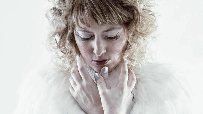 A performer stands against a white background with eyes closed, wearing a blue dress with a white feathered collar.