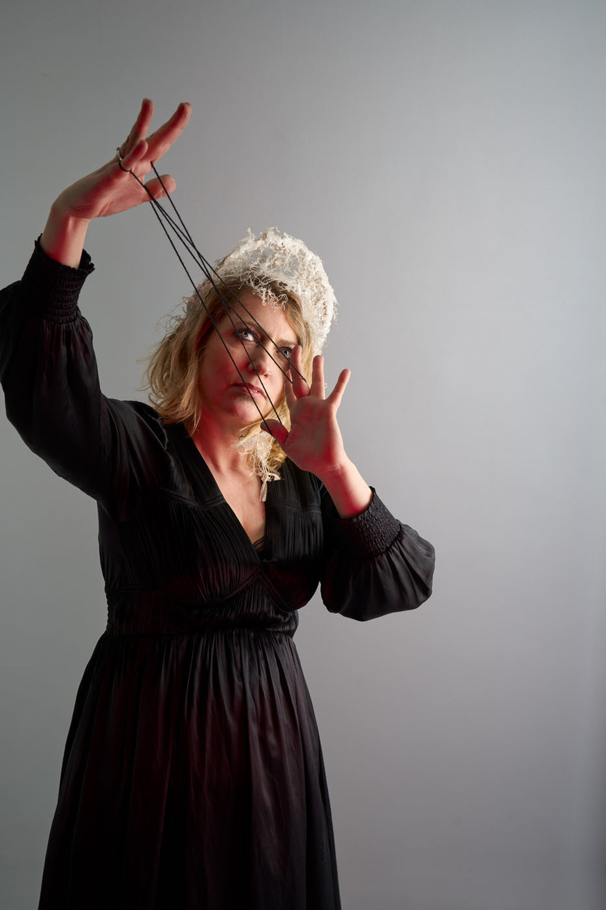 A performer wearing a black dress & white headpiece. Holding black strings that make diagonal lines on their face.