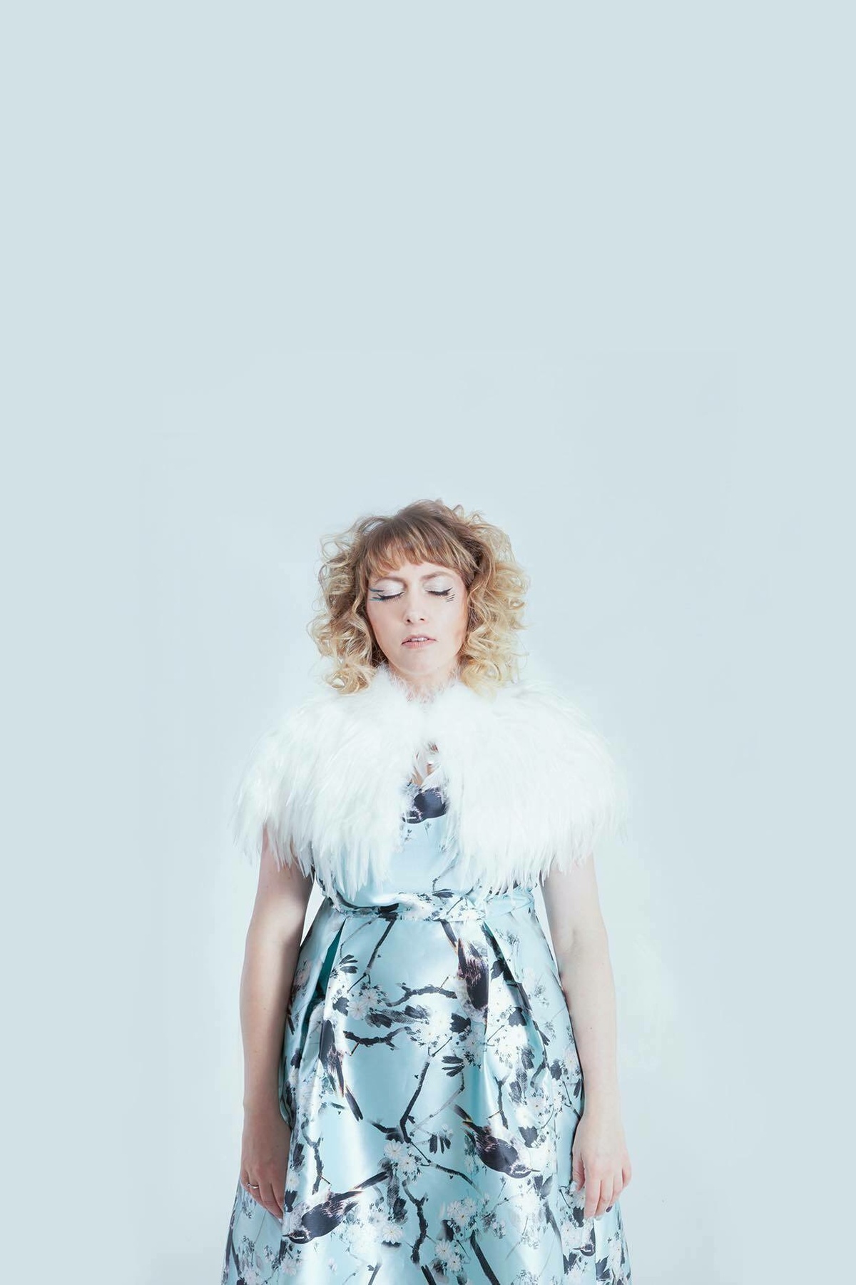 A performer stands against a white background with eyes closed, wearing a blue dress with a white feathered collar.