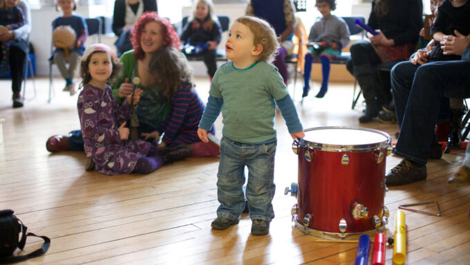 A photograph of young children with their parents and carers, centred is a smiling child with their hand on a drum
