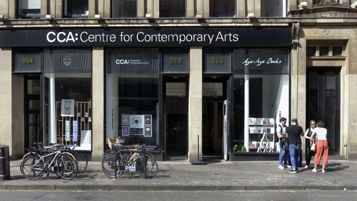 A photo of the front doors and windows of the CCA building with bicycle racks and a small group of people on a sunny day