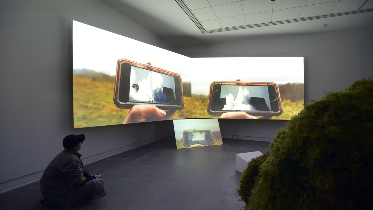 A photo of a dark exhibition. A person sits on the floor looking at a three channel projection in front of a mossy chair