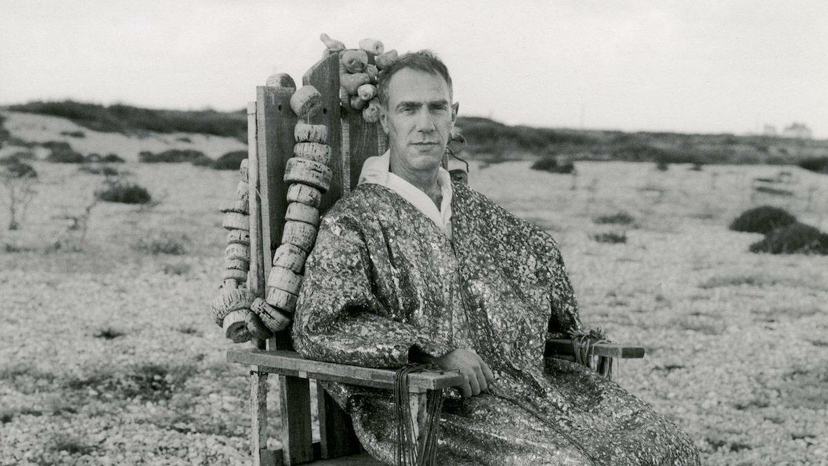 A black and white photograph of the artist Derek Jarman, kind and stoic, in a sequined robe on a driftwood throne.