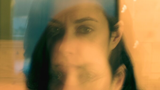 A double-exposure of a face. They have long dark hair which is tucked behind their left ear and are wearing mascara.