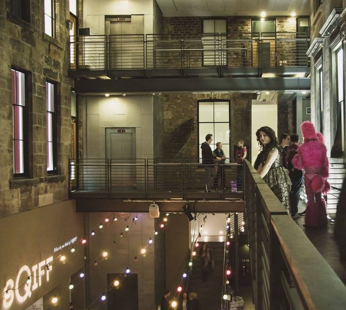 A group of people gather in CCA at night, one looks at the camera, another is in a pink suit