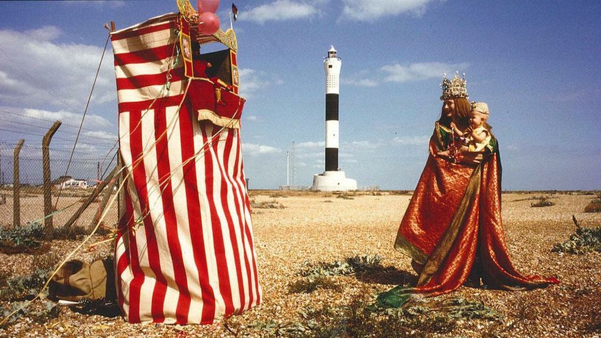 Tilda Swinton holds a child in her arms as they approach a Punch and Judy stand on a shingle beach.