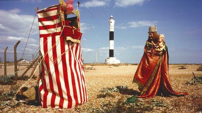 Tilda Swinton holds a child in her arms as they approach a Punch and Judy stand on a shingle beach.
