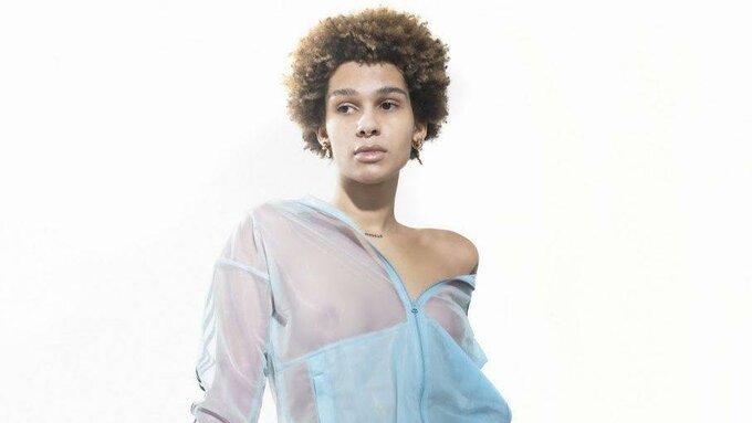 TAAHLIAH is sitting against a white backdrop wearing a sheer blue tracksuit. She is looking off camera.