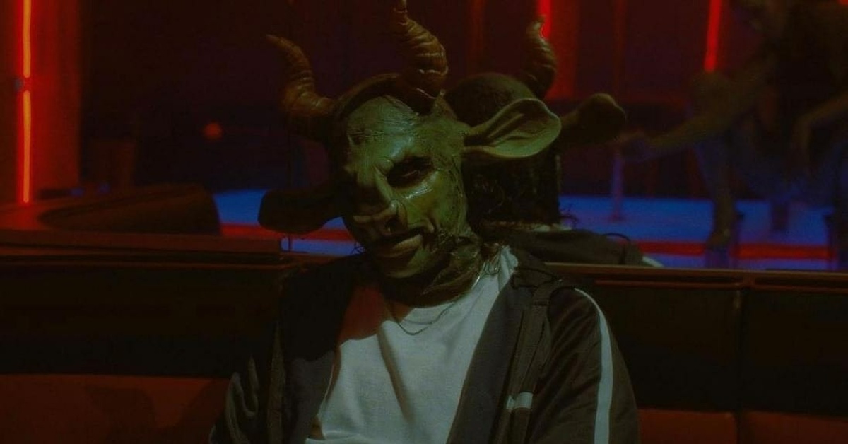 SHREK 666 is sitting in a dark room wearing a green prosthetic mask with a snout nose, shrek style ears and horns