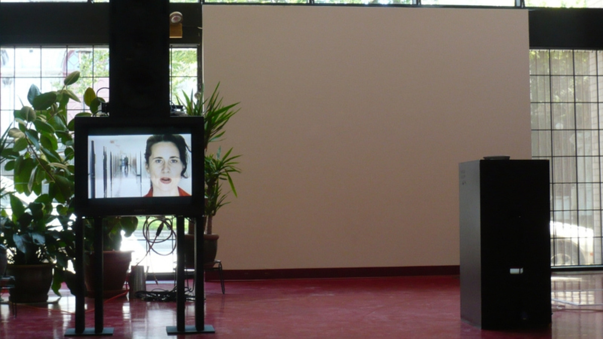 A room containing a television and a large white projection screen with plants. A person wearing red is on TV.