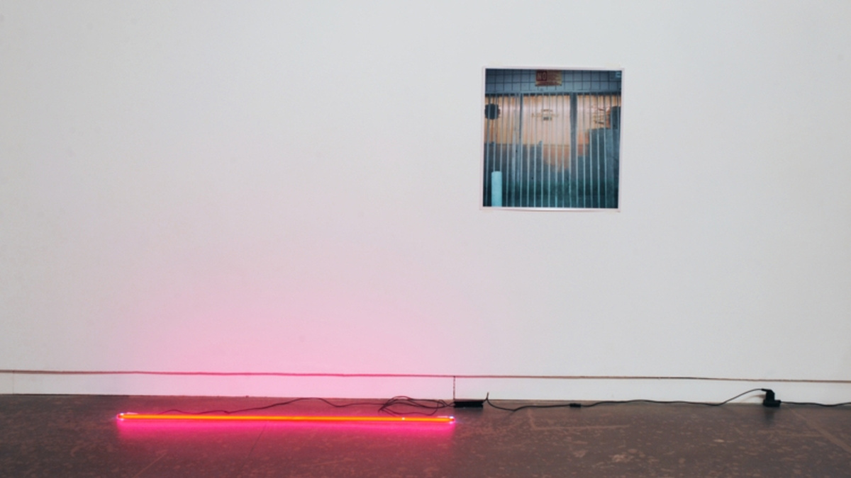 A picture is hung on a white wall, a pink neon lighting strip is on the floor.