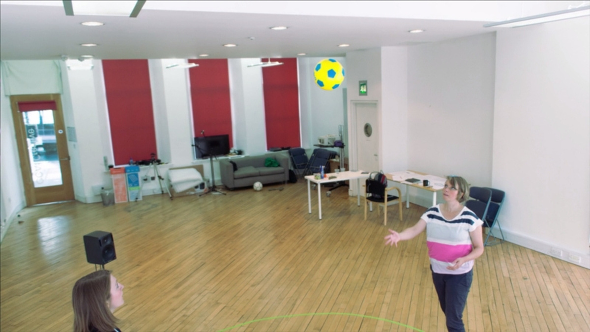 Two people stand in a room with a wooden floor. They are passing a bright yellow football to each other.