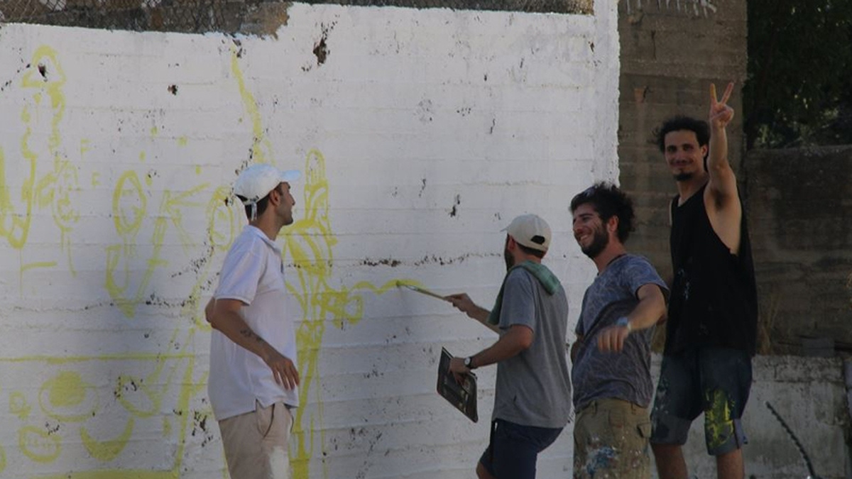 Four people paint a wall in Palestine, one is making the peace sign with his fingers to the camera.