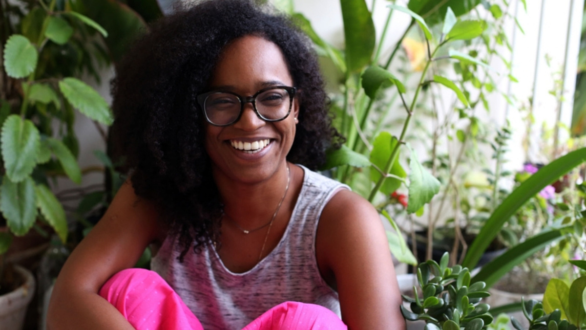 A portrait of Nicole Smythe-Johnson, she is sitting with her knees up and smiling. She is surrounded by green plants.