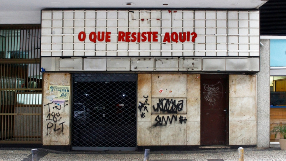 A run-down cinema with grafitti and shutters. The board reads O Que Resiste Aqui? Translation: What resists here?