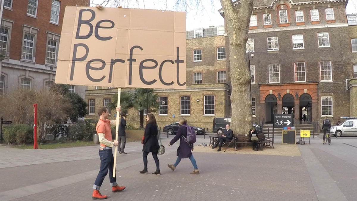A person stands in a public square holding a large cardboard sign reading "Be Perfect".