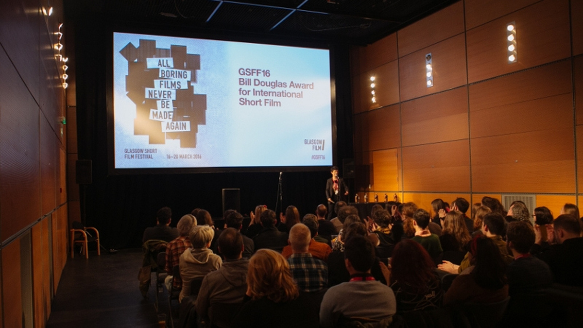 A photograph of the CCA cinema, there is a large audience and a projected screen with the GSFF logo.
